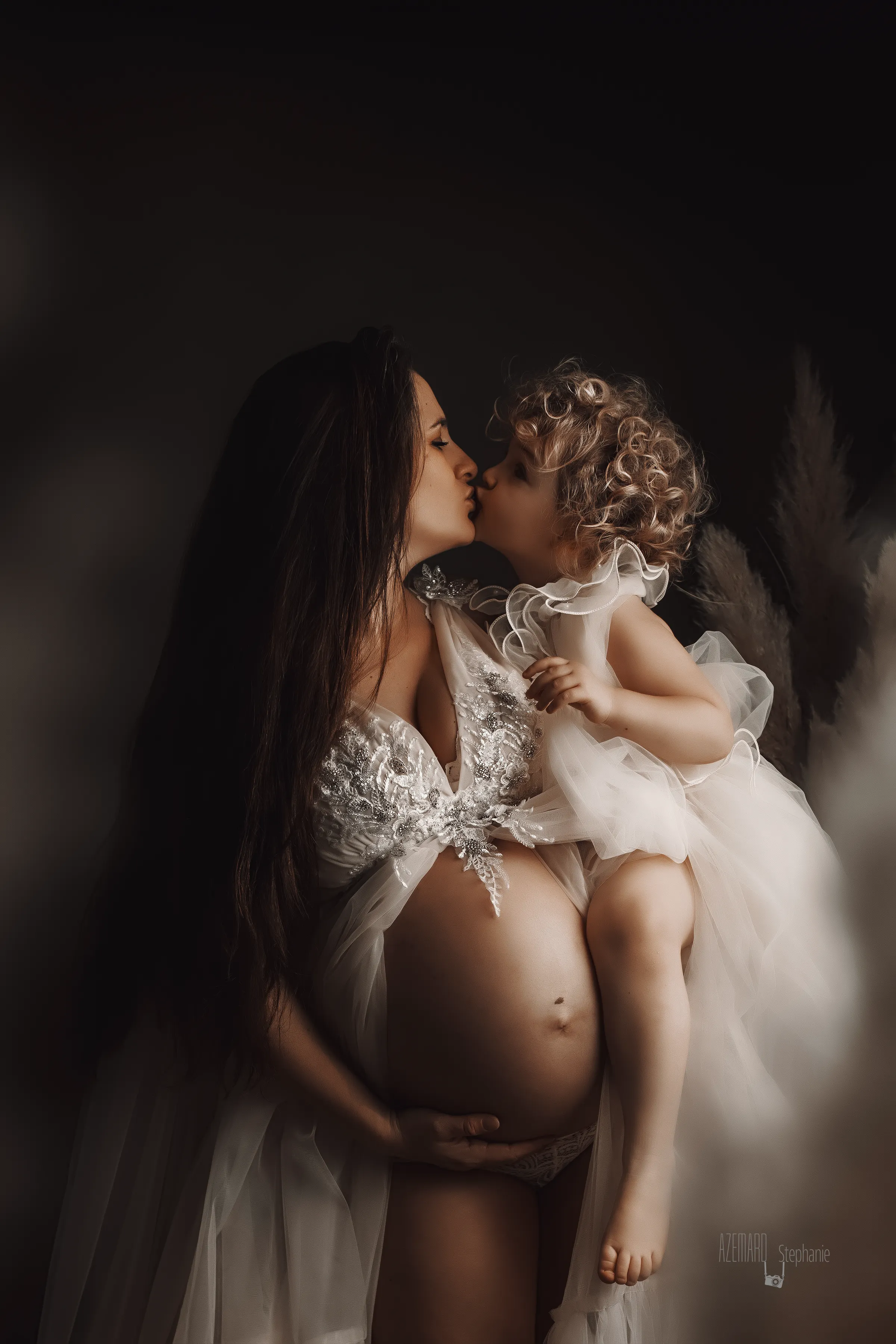 Photo grossesse maman enceinte embrassant sa fille habillée en ange en studio fond sombre, photographe maternité Montpellier Stéphanie Azémard