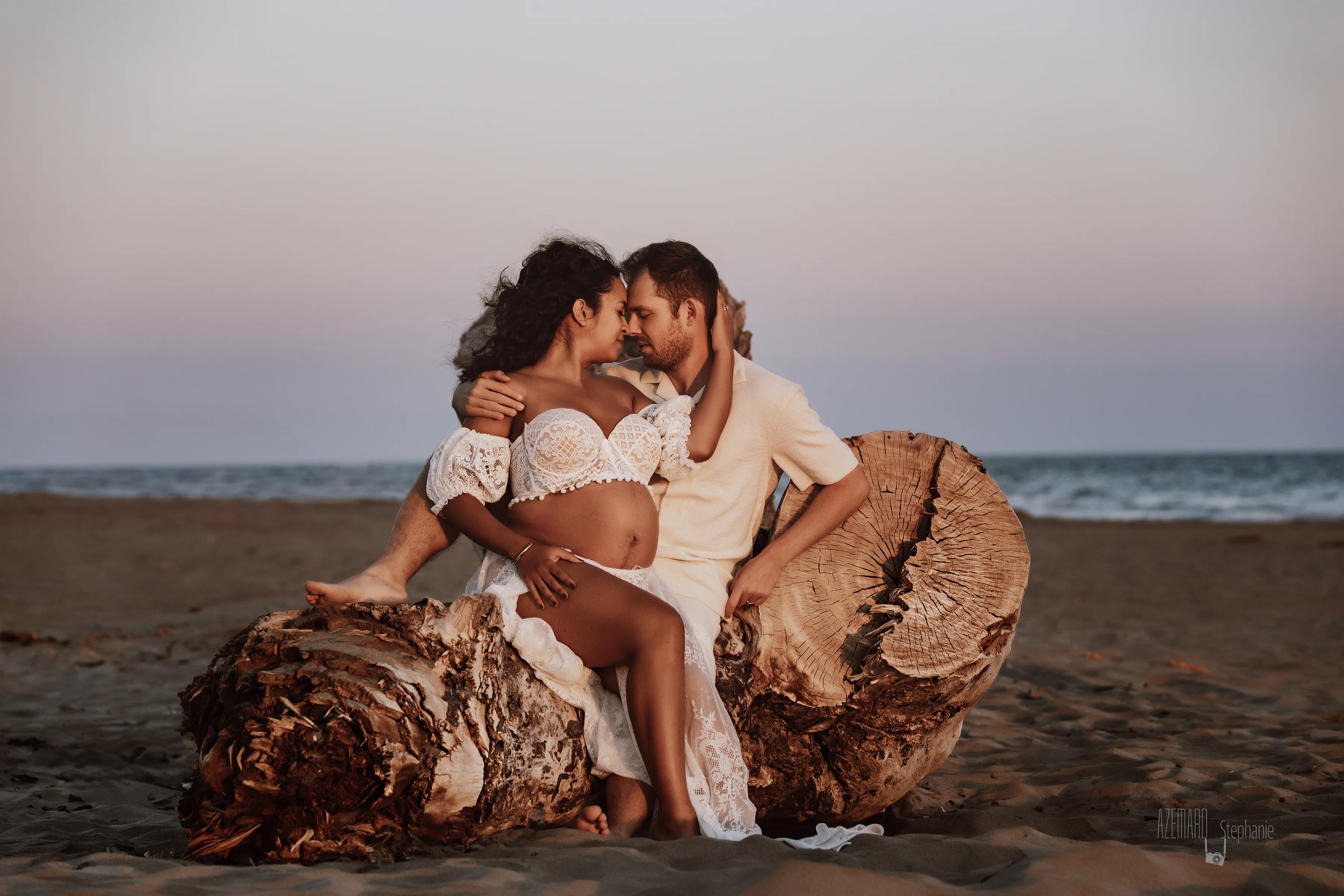 Photo grossesse couple assis sur tronc bois flottée plage au coucher de soleil, future maman dentelle blanche, photographe maternité Montpellier Stéphanie Azémard